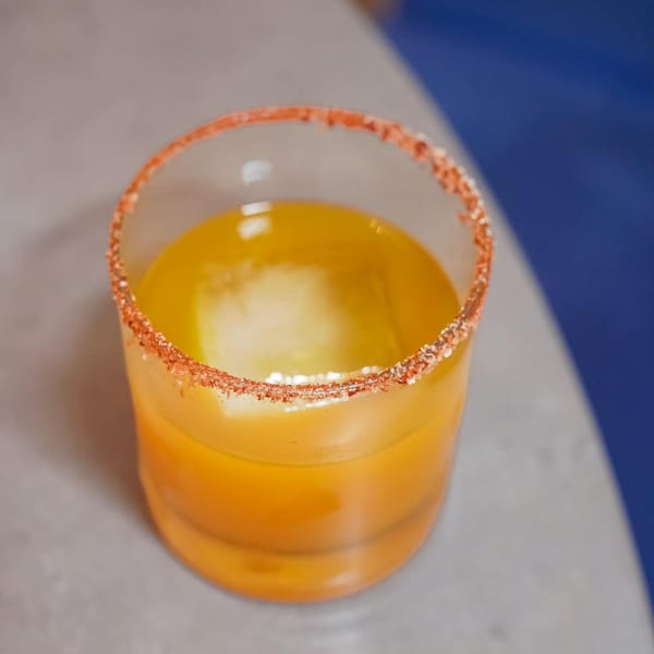 A vibrant orange cocktail with a chili-salt rim and a large ice cube, on a gray and blue surface.
