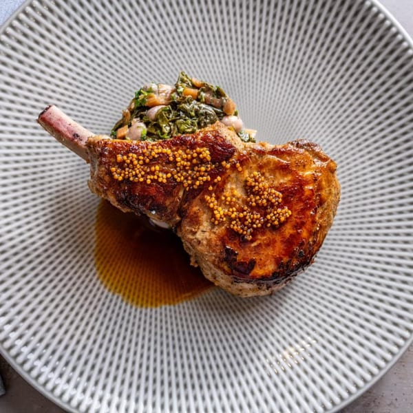 Grilled pork chop with whole grain mustard, braised greens, and a rich sauce on a patterned plate.