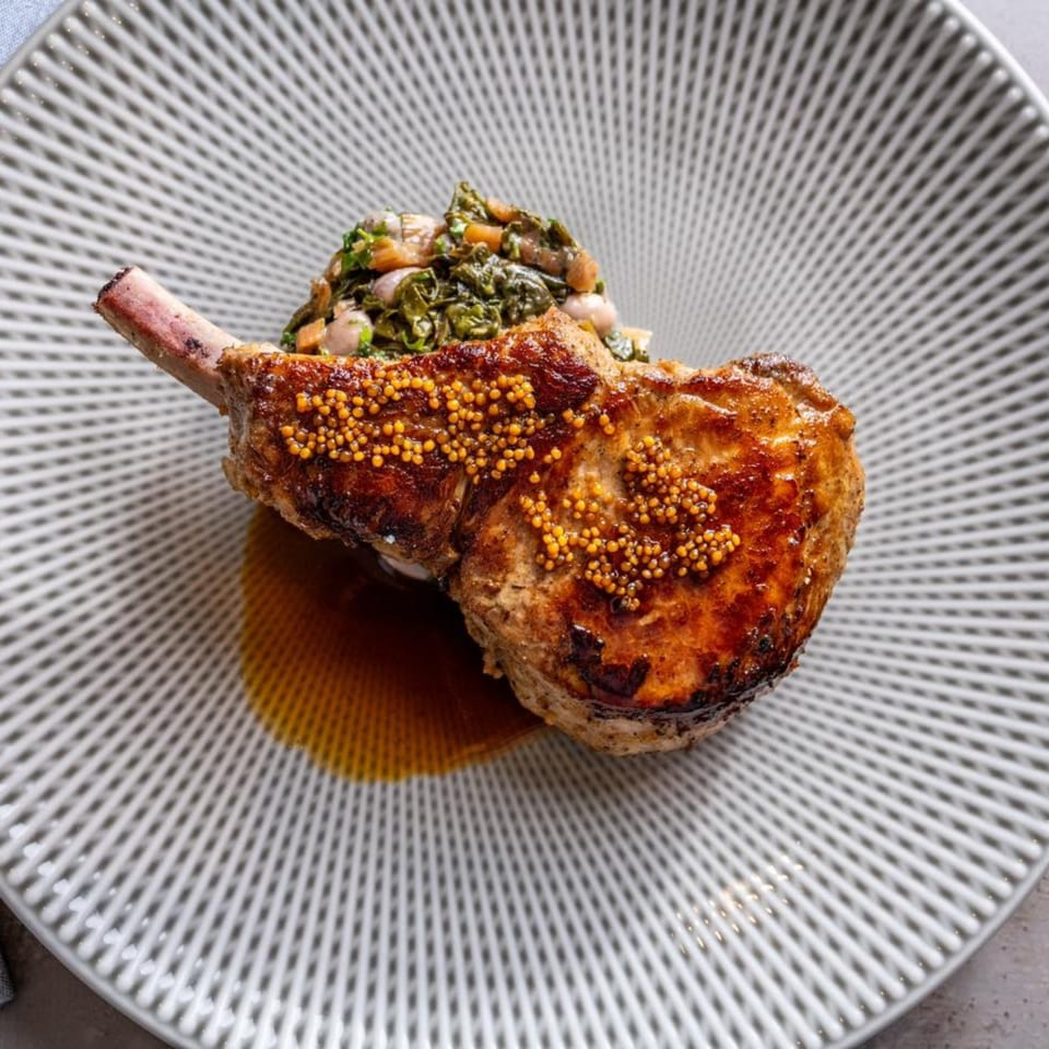 Grilled pork chop with whole grain mustard, braised greens, and a rich sauce on a patterned plate.
