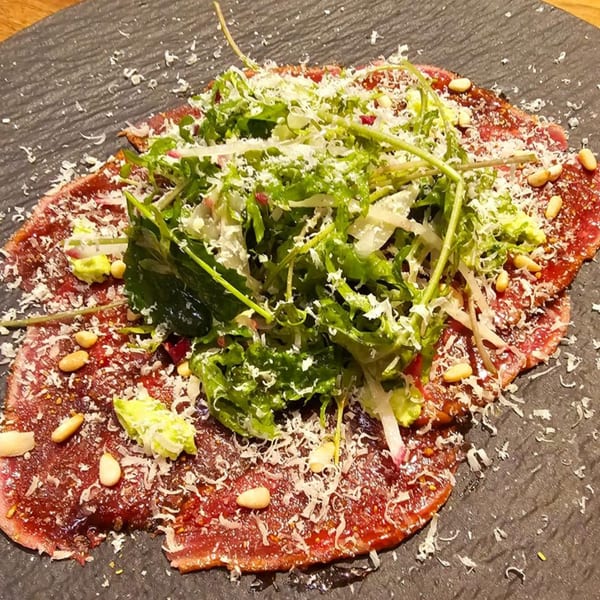 Carpaccio with arugula, pine nuts, and grated cheese on a dark plate.