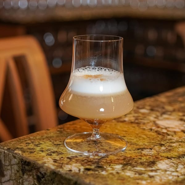 A cocktail with a foamy white top layer sits on a speckled brown bar counter.