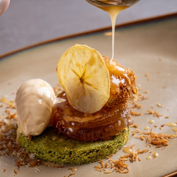 Dessert with green cake, caramel, ice cream, banana chip, and honey drizzle on a plate.